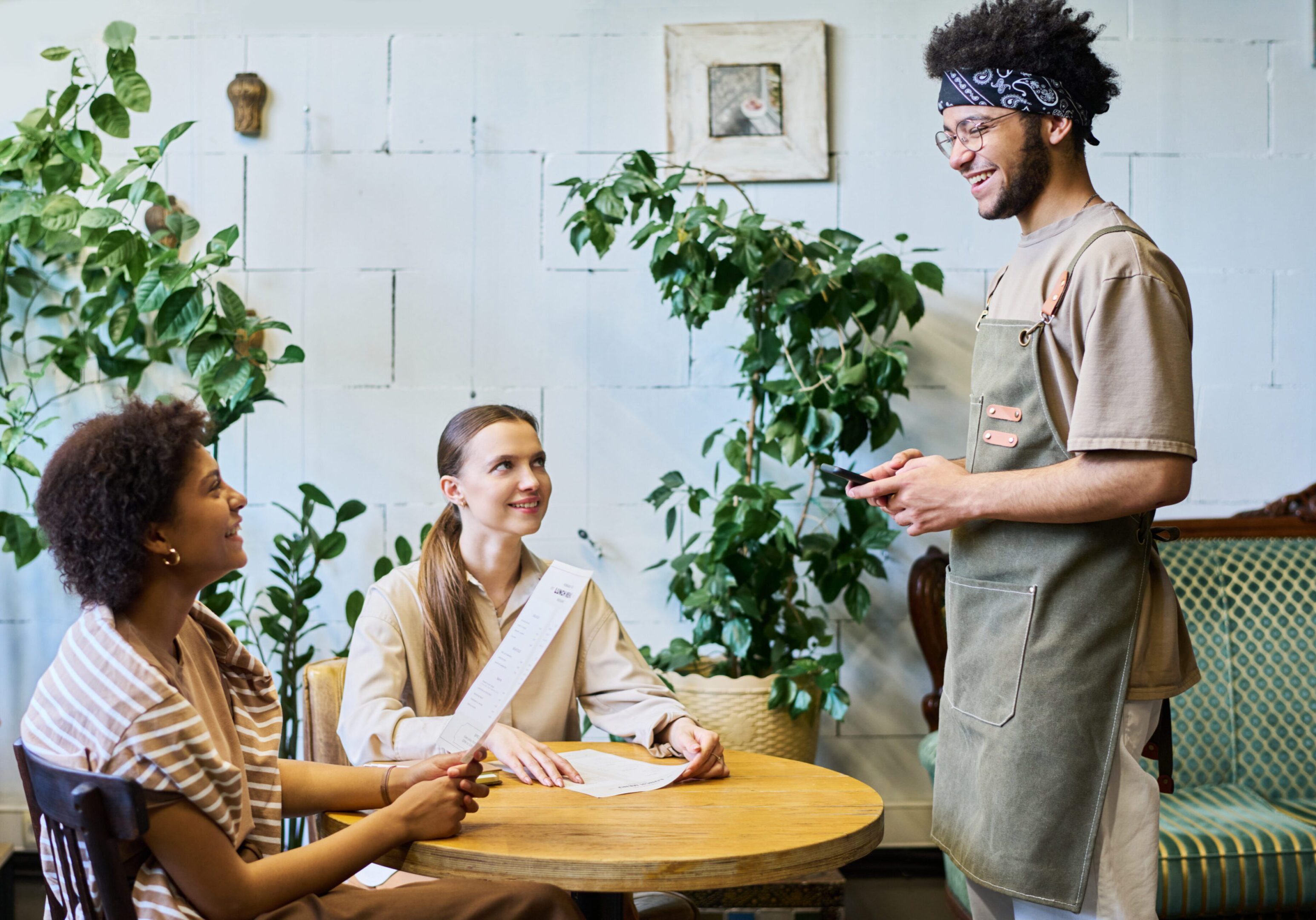 Two intercultural girls with menu looking at smiling waiter in workwear standing by their table and waiting for their orders in cozy cafe