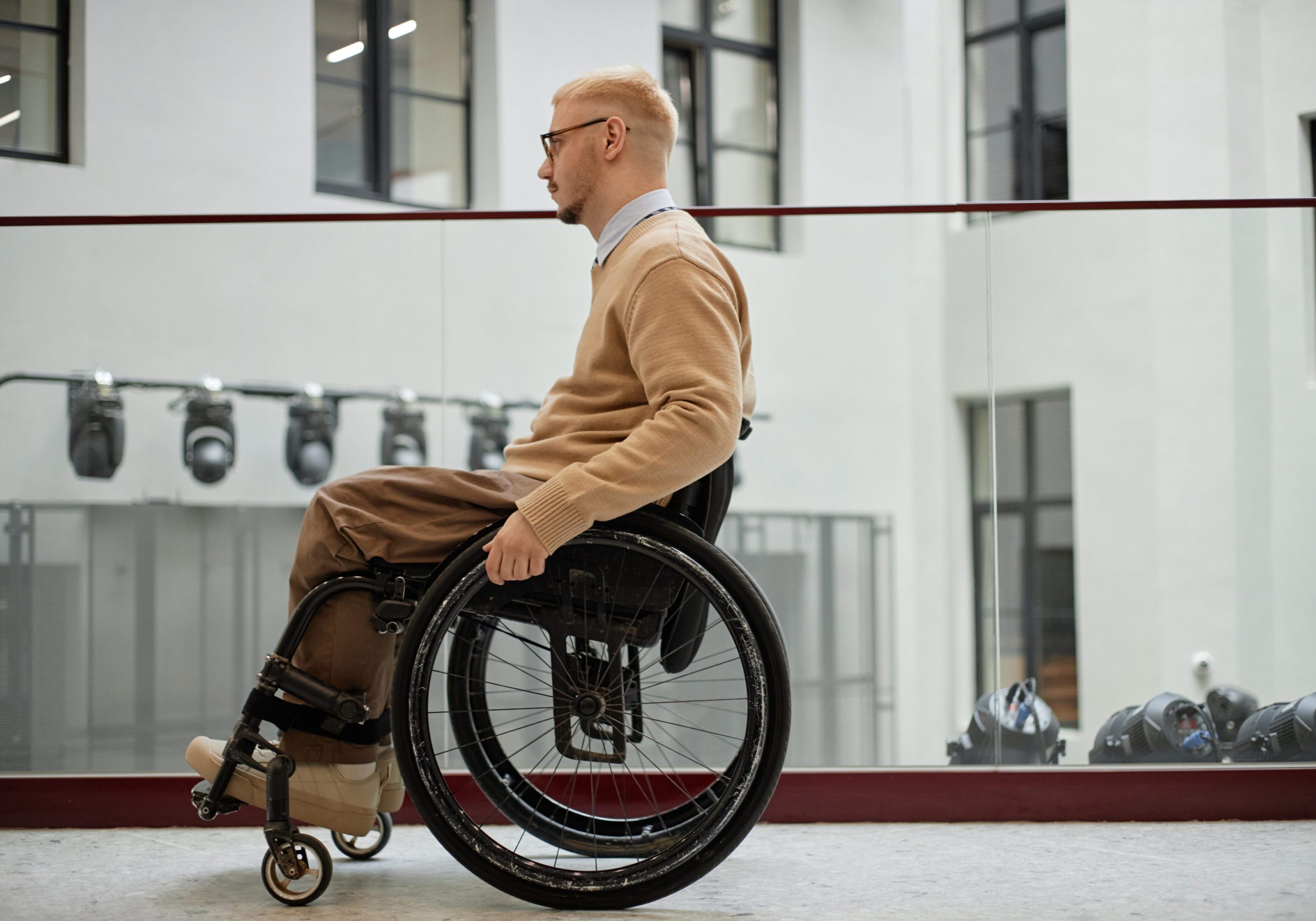 Caucasian young adult man with disability using wheelchair indoors, sitting in profile view near glass railing, wearing eyeglasses